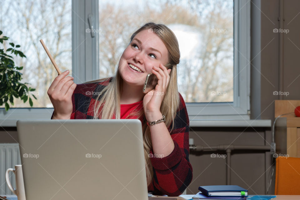 a smiling blonde woman sitting at a laptop and talking over a smartphone
