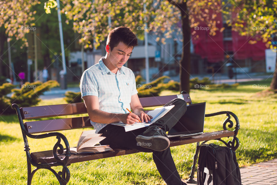 Student working on a laptop using books and notes sitting on a bench in a park. Young boy wearing a blue shirt and dark jeans