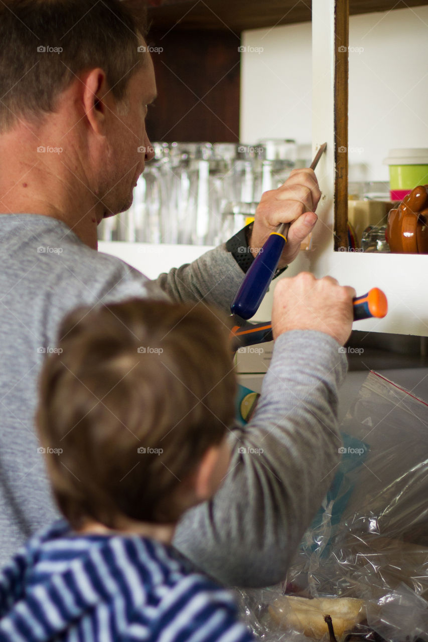 DIY at home - dad and his little helper boy working to repair and improve the kitchen cupboards. Image of dad and son while dad uses tool to repair hinges.