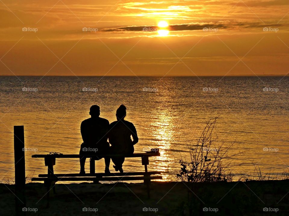 
Living in harmony - life is good. A romantic couple sitting on a picnic table spend time together alone, away from family and friends watching a magnificent sunset descending over the shimmering bay. 🎊🎉