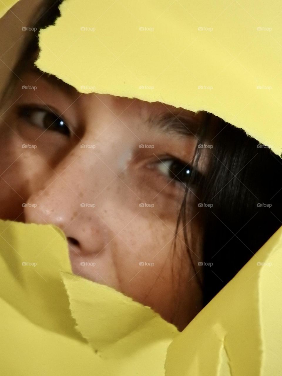 Face skin in the forties of a woman with freckles and brown hair seen through a broken yellow paper.