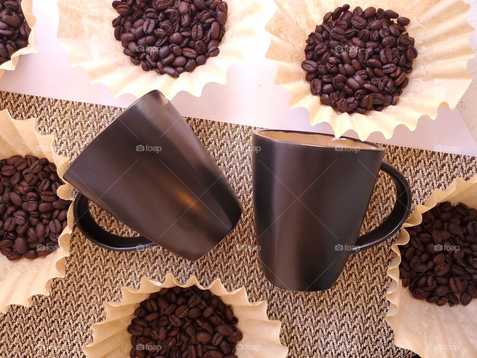 Coffee beans displayed in coffee filters surrounding two black coffee cups on partial white and partial beige surface.