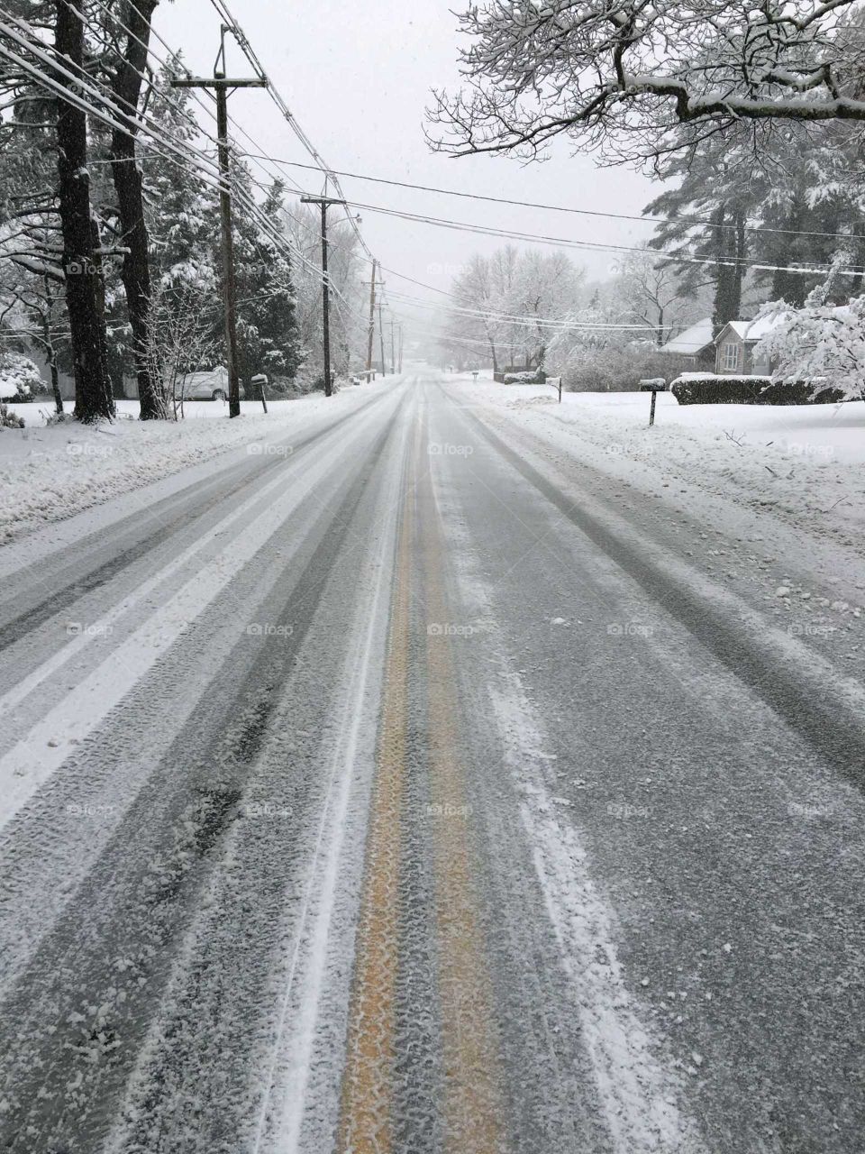 Snow covered rural road, straight road, no cars, double yellow lines visible through icy snow. No people are out shoveling during snowstorm. Winter weather, snow, pic in middle of the road. Almost monochromatic B & W pic! Snow covered street.