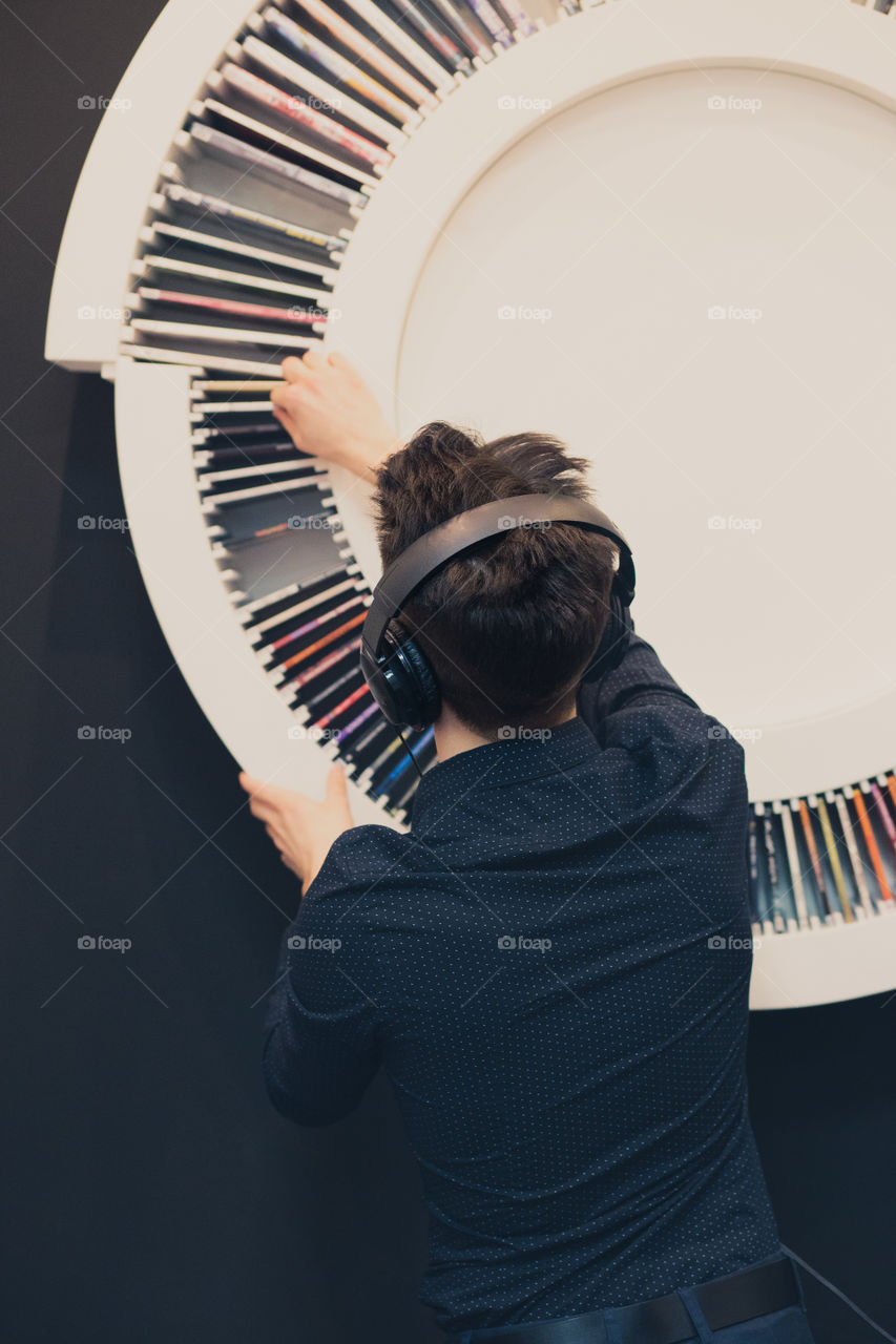 Young man listening to music through headphones standing next to bookshelf with recordings