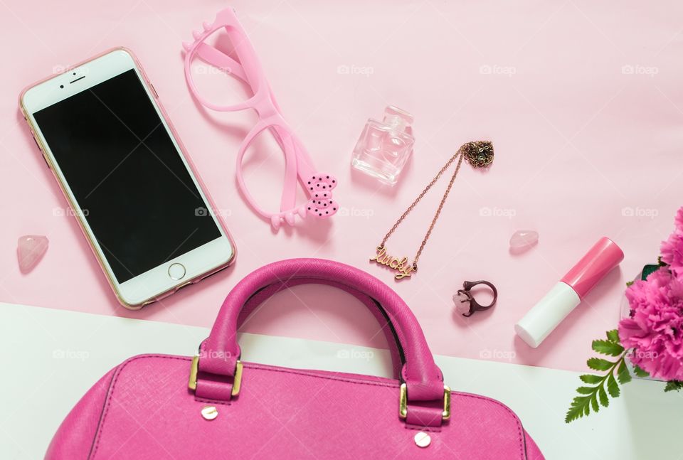 Flat lay of items - mobile device, glasses, perfume, rings, jewelry, lipstick, and flower surrounding a pink bag