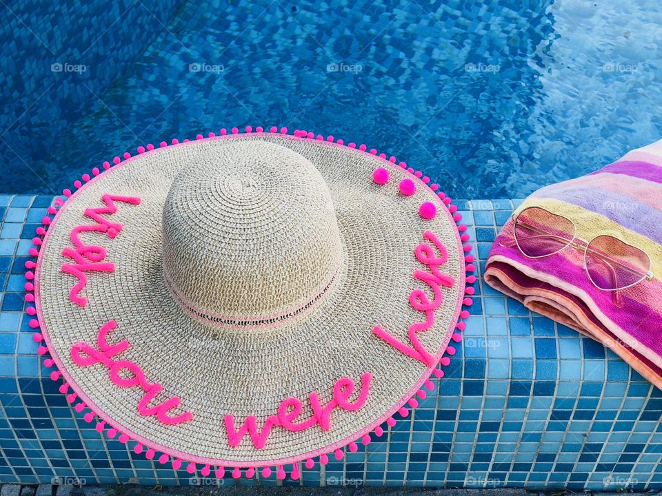 Summer hat with the words “wish you were here”,sunglasses and a pink towel on the edge of a pool with turquoise water