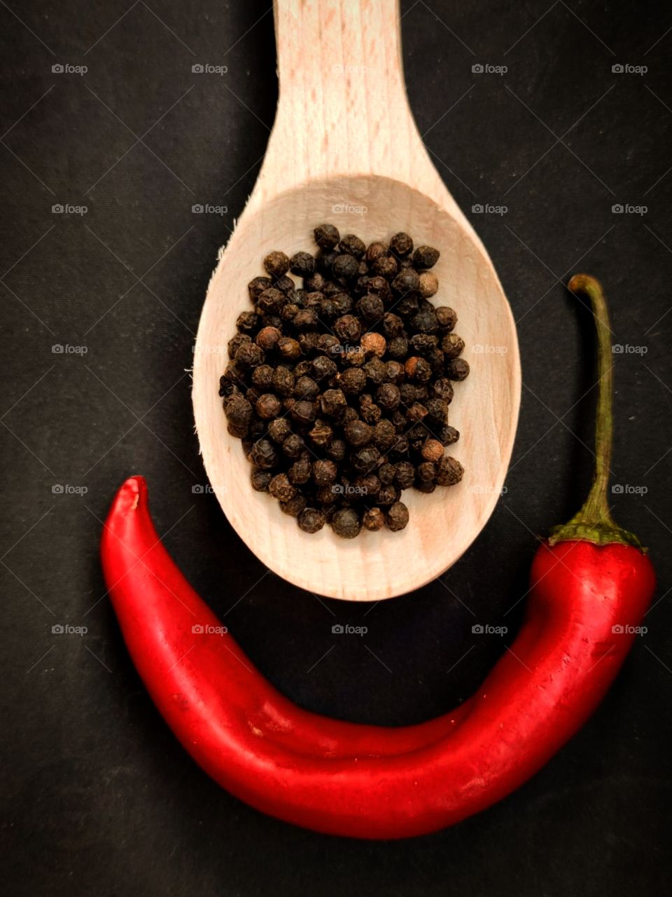 Spices on a black background.  There are black peppercorns in a wooden spoon.  Red chili peppers under the spoon