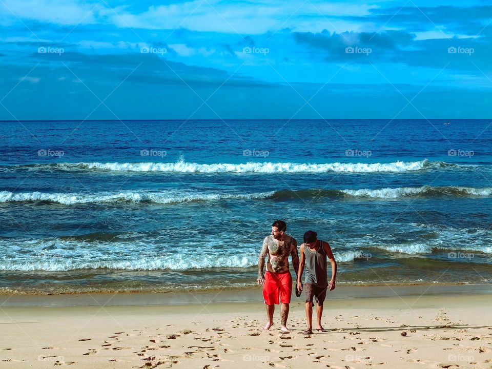 Two men walking on the beach, at sunrise, with a Beautiful Blue sky and ocean waters behind them.
Tattooed men and his friend pn a Summer holiday tropical vacation.