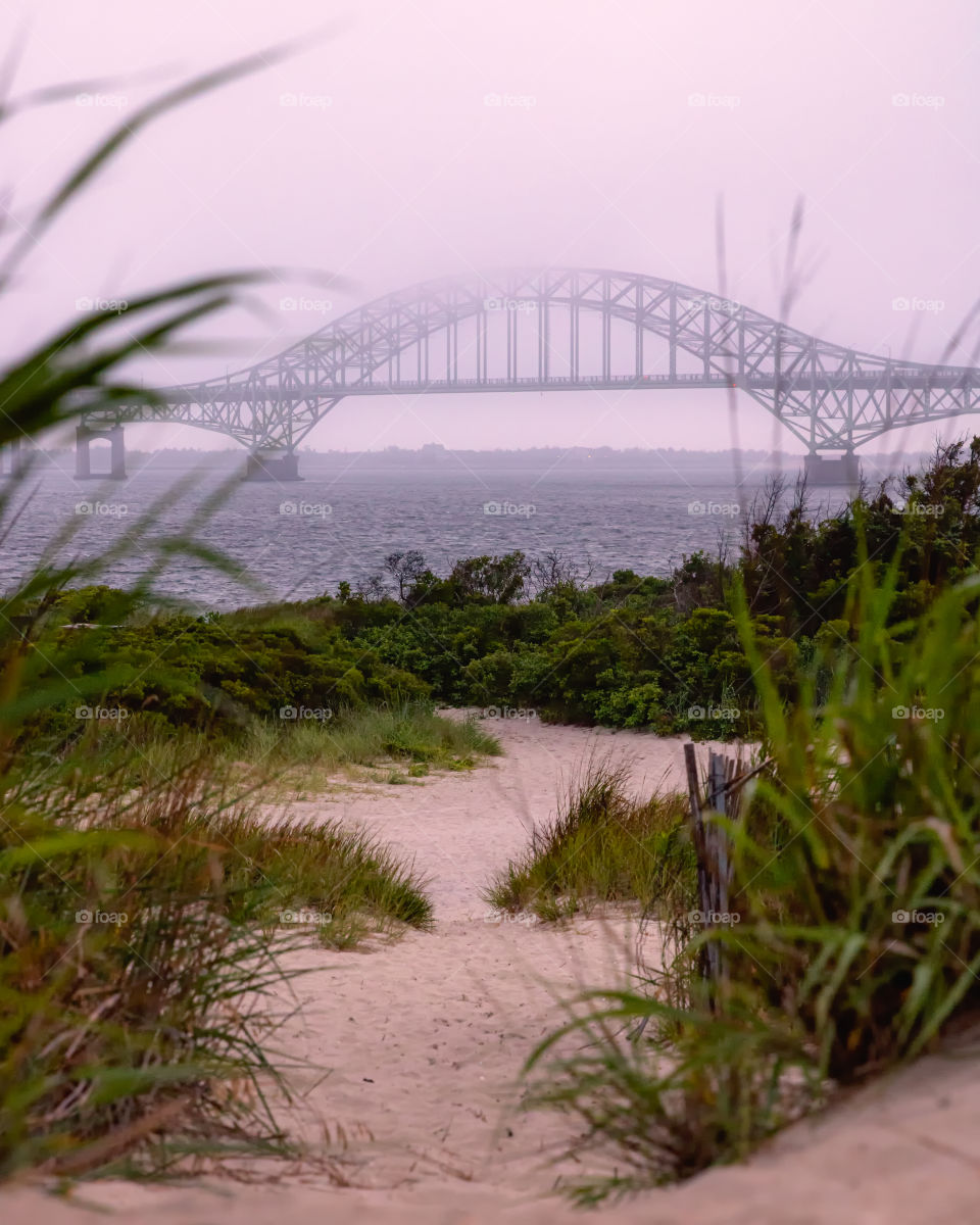 Fog and mist ccoming in off the ocean, engulfing an arched bridge and moving towards a sandy grassy coastal beach 