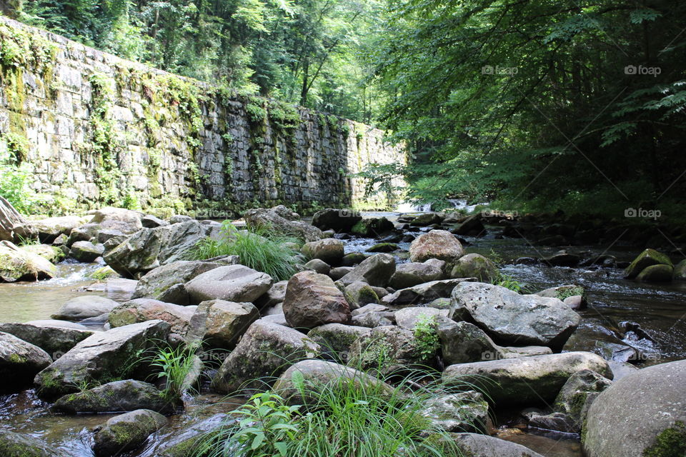 Giant stones pave the way down a creek in a park. The summer sunshine lights of the stone wall off the left