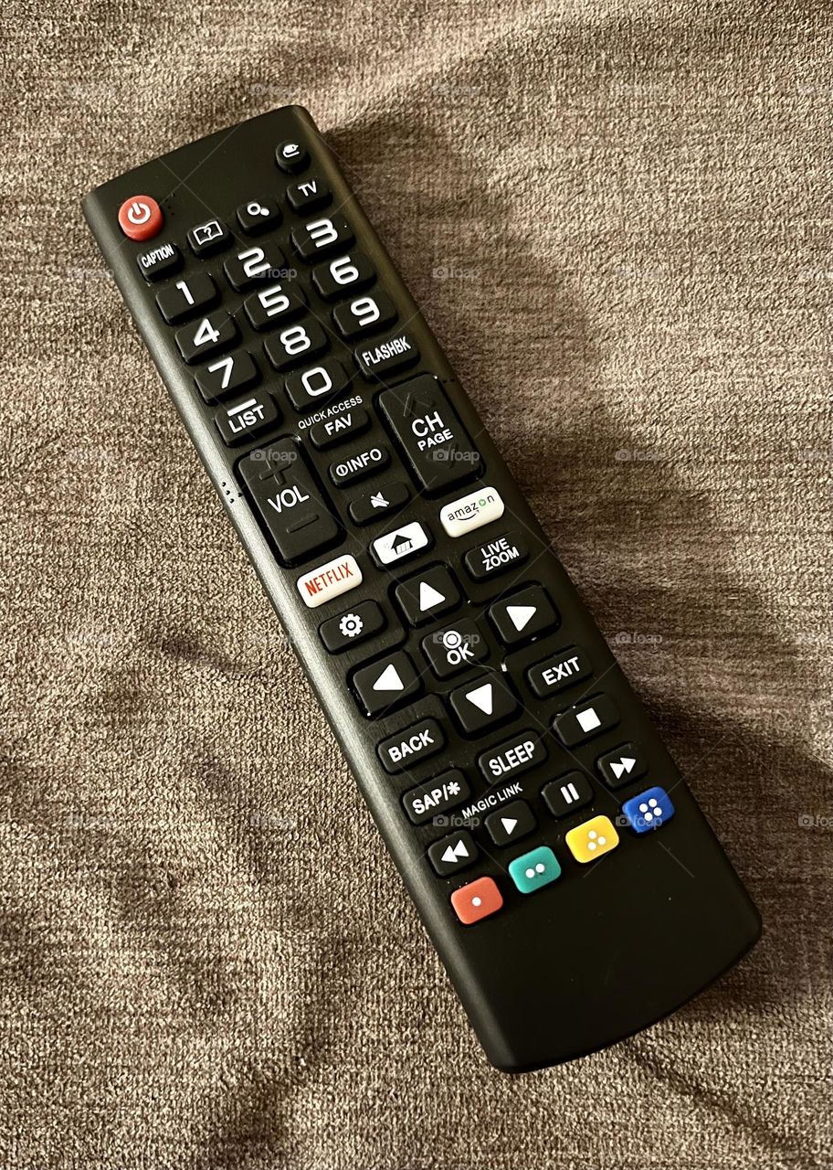 Black rectangle tv remote control on the couch in the living room of the home.  Used to work the tv, change the challenge, power off and on, volume controls with colored buttons to push.