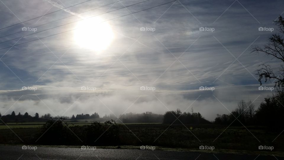 bright sun behind a thin blanket of clouds above a landscape of field and trees with a misty background