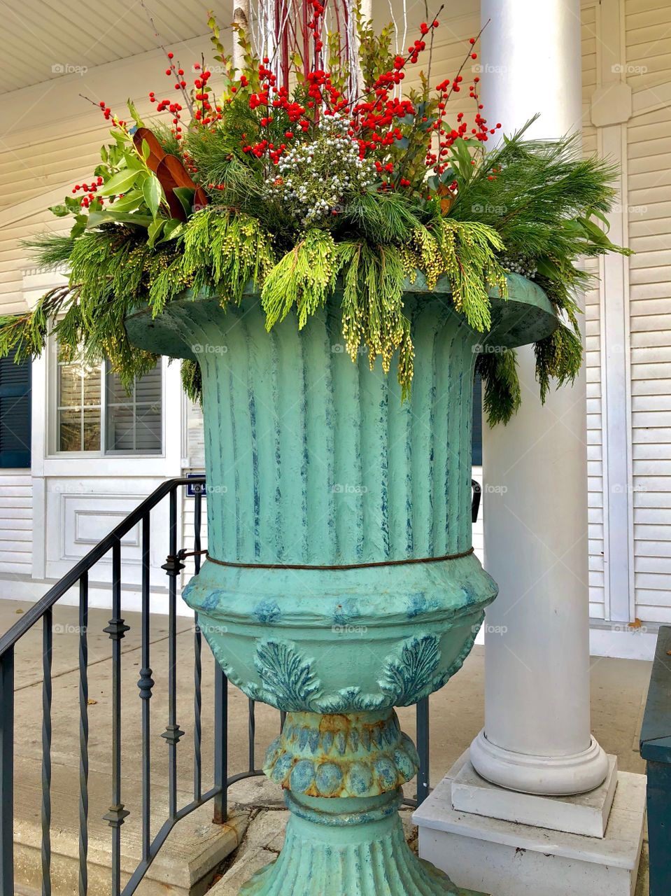 "Holiday Decor" - A large green sculpted ceramic flower pot with decorative trim is filled with greenery, flowers & red berries to welcome the Winter holiday season. It stands at top of front steps by a white concrete column outside a white building.