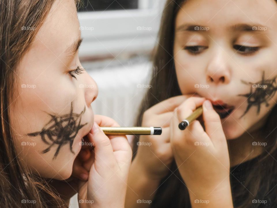 Portrait of beautiful little caucasian girl doing Halloween holiday makeup with pencil and reflecting in the mirror, vtd side close-up.