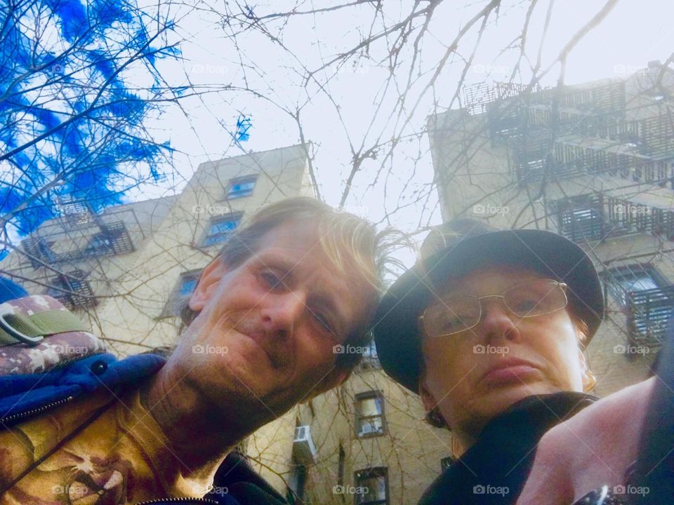 In front of an Upper E Side apartment building beneath a beautiful blue sky w perfectl little white cumulus clouds in it longtime partners in life, love & everything, “Petra Schiller” & “Timothy Trespas” are smiling. 2020. Hypnotic Productions