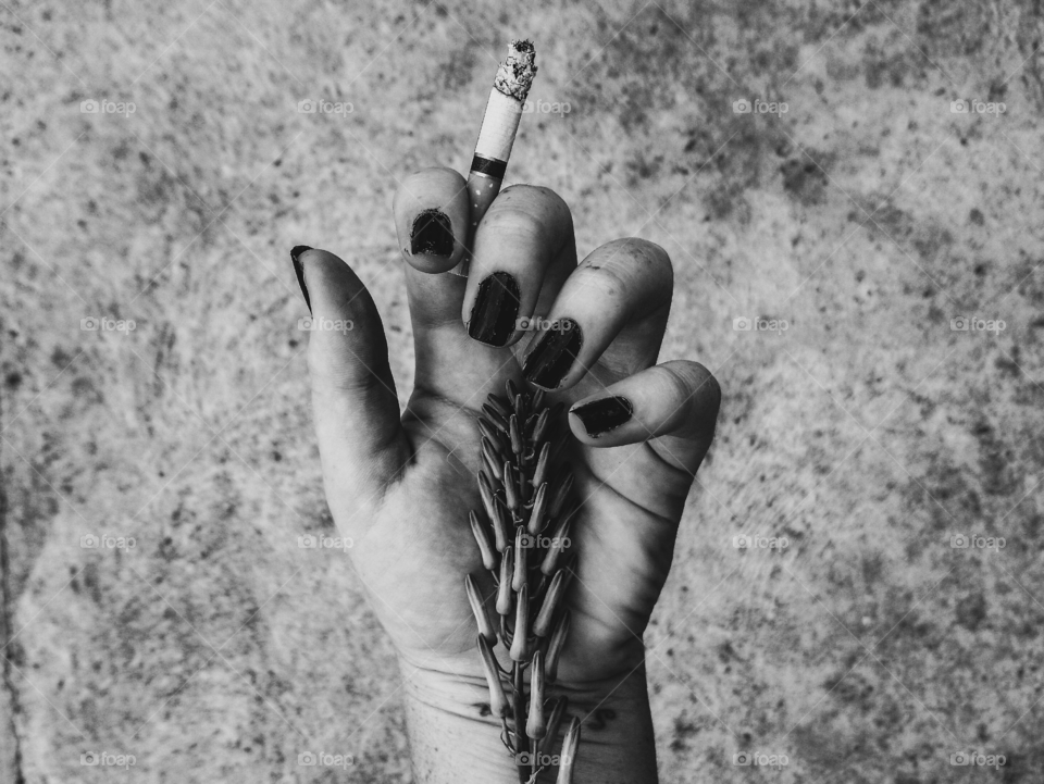 Women's hand holding cigarette with plant and painted black nails, depicting addiction