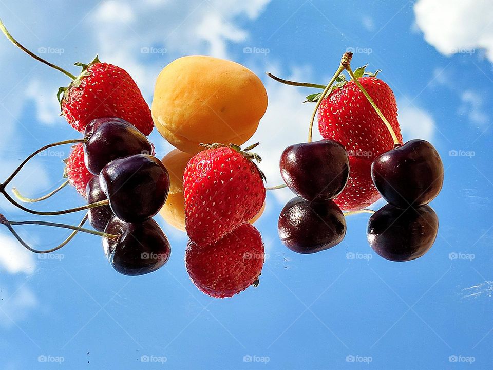 Summer fruit symphony. Fruits lie on a mirror surface: red strawberries, black cherries and yellow apricot. Fruits and a blue sky with white clouds are reflected from the mirror surface.