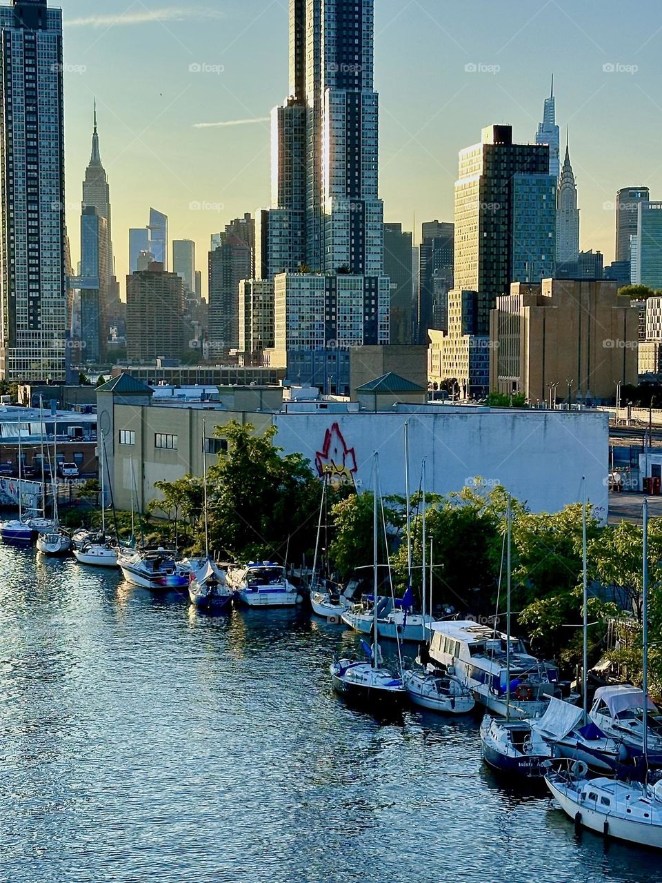 This is a closeup of the LIC shore with its many boats seen from the “Pulaski Bridge” that connects “Greenpoint”, Bklyn to LIC, Queens. “Manhattan” can be seen in the distance including the “Empire State Building”. 2024. Hypnotic Productions