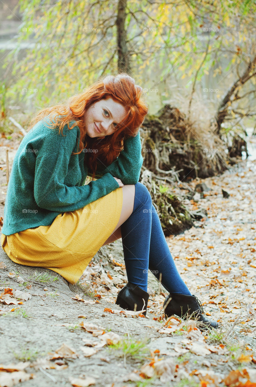 Portrait of young happy redhead woman sitting and enjoying near autumn lake in october.