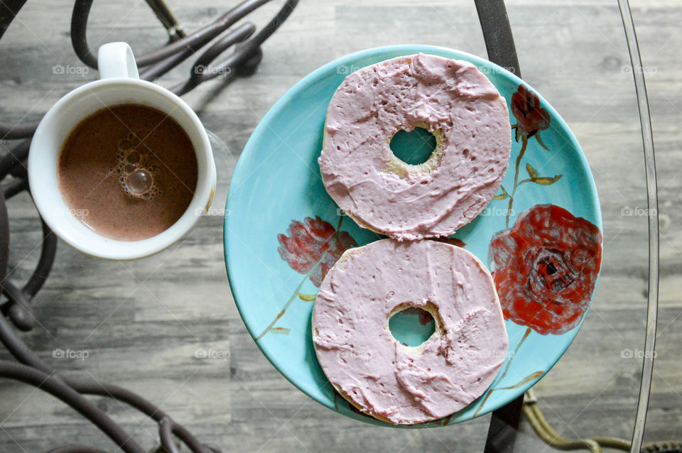 Colorful plate of purple berry cream cheese on a bagel next to a mug of chocolate milk on a glass table