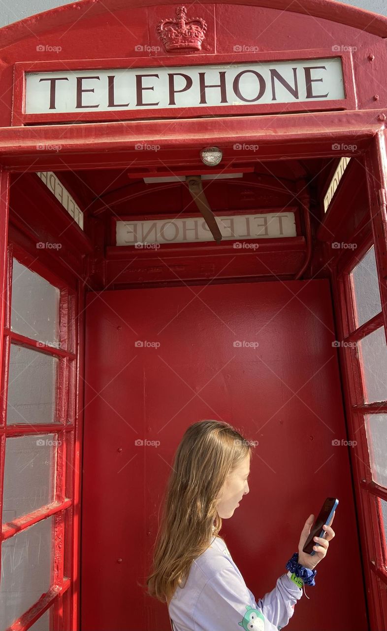 A girl looking at her cellphone while in an old English style red phone booth. 