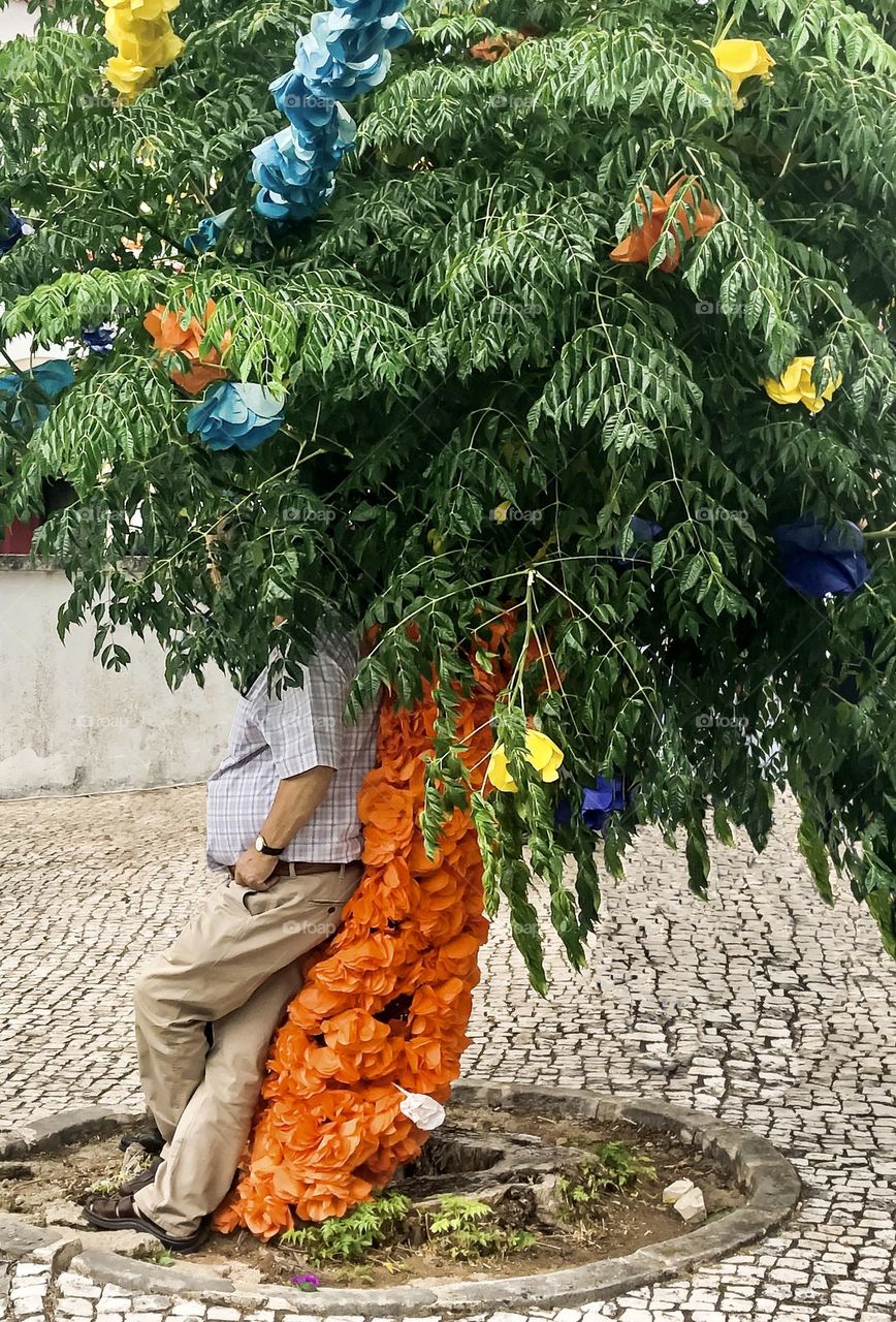 Man man rests against a tree decorated with coloured tissue paper, enjoying the shade