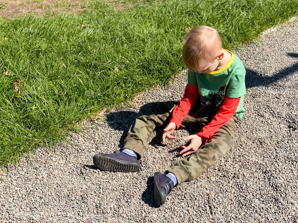 the boy sits on the walkway and plays with gravel, green grass