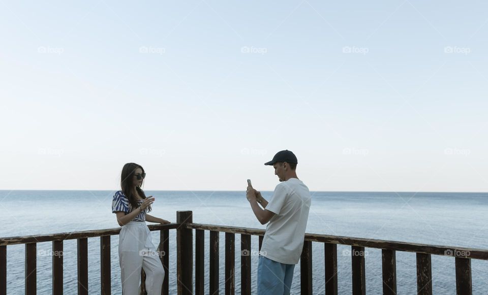 One young handsome Caucasian guy takes a photo of his girlfriend on a smartphone while standing on a wooden bridge against the backdrop of the sea on a summer evening, close-up side view.