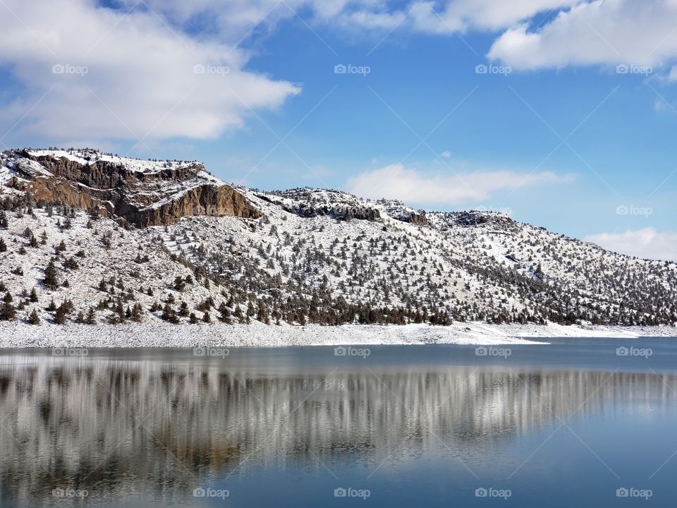 Snow covered hills, trees, clouds, and a bright blue sky reflect in the partially iced over Prineville Reservoir on a sunny winter day in Central Oregon. 