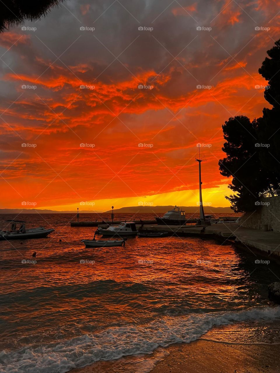 Amazing red sunset scene over the Mediterranean Sea with dramatic reflection in the lightly waving water surface near the pier with few boats