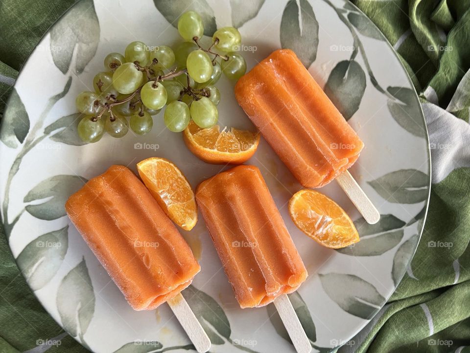 Three orange popsicles with Clementine slices and green grapes on a leaf-printed plate set on a green and beige  tablecloth 