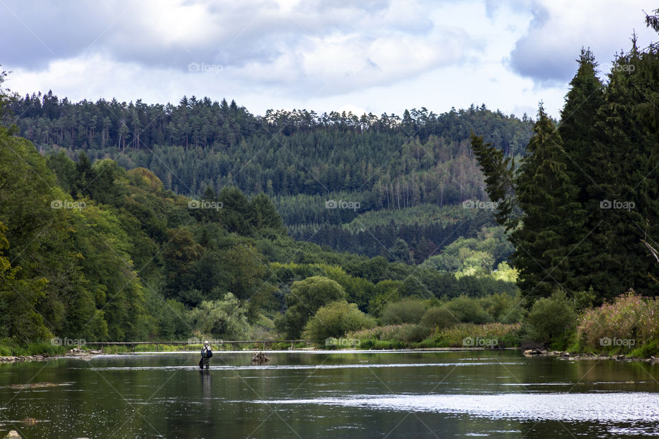 A landscape shot of a river in the middle of the woods in the Belgium ardennes in la fayette. there is a fisherman in the river doing some fly fishing.