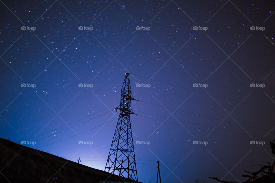 The tower of the power line stands on the mountain at night, against the background of the stars.