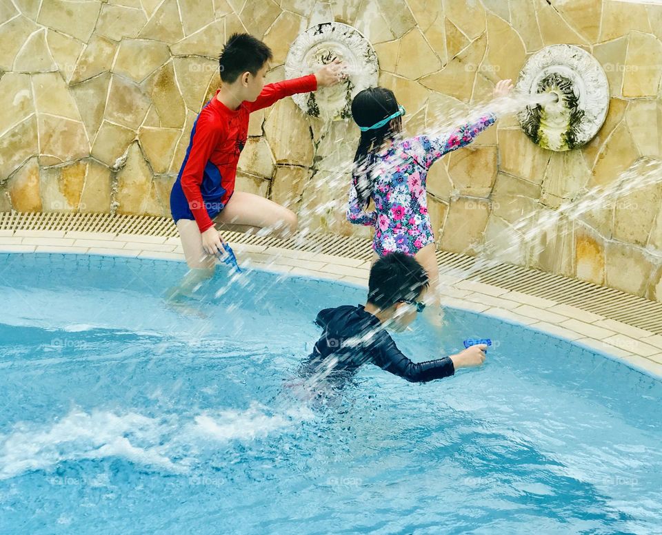 Children love to play and enjoy at swimming pool in summer , here three children playing under flowing water (fountain) and water sprinklers in their hands . Totally it’s a fun time for everyone.