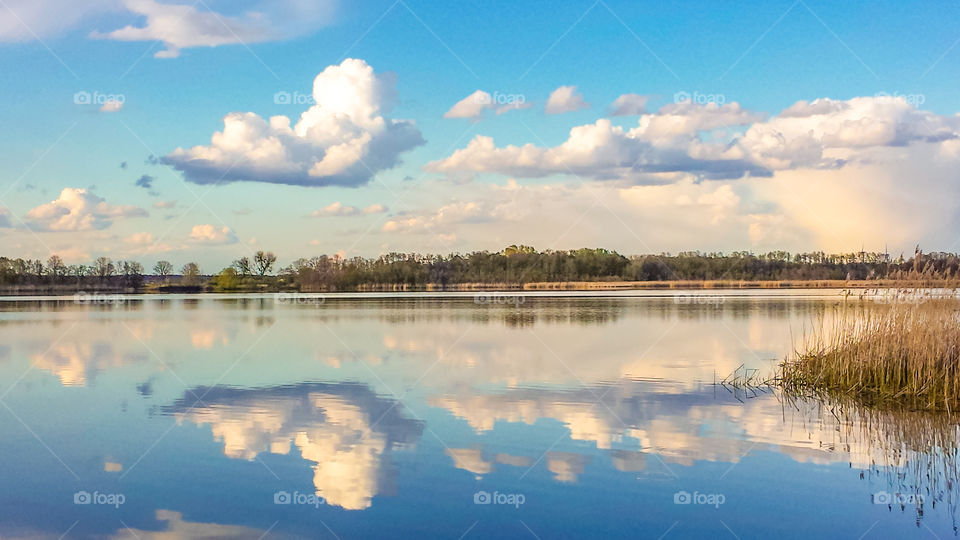 Der Geronsee  bei schönem Wetter, die Wolken spiegeln sich im Wasser. Im Hintergrund ist bewaldetes Ufer, im Vordergrund etwas Schilf.