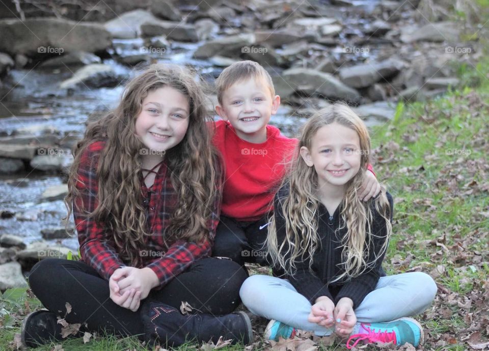 A brother affectionately hugging his two sisters in front of a beautiful mountain stream. 