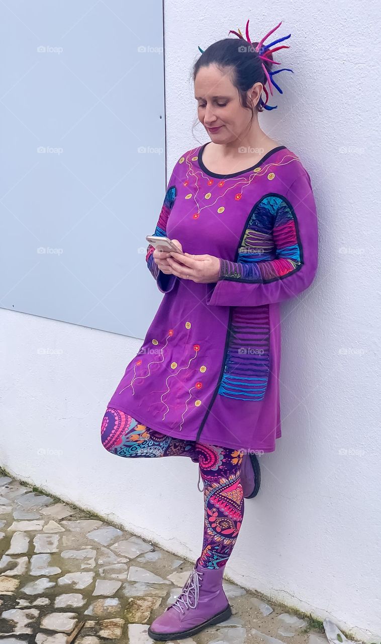 Woman in purple dress and colourful leggings leans casually against a wall browsing on her phone