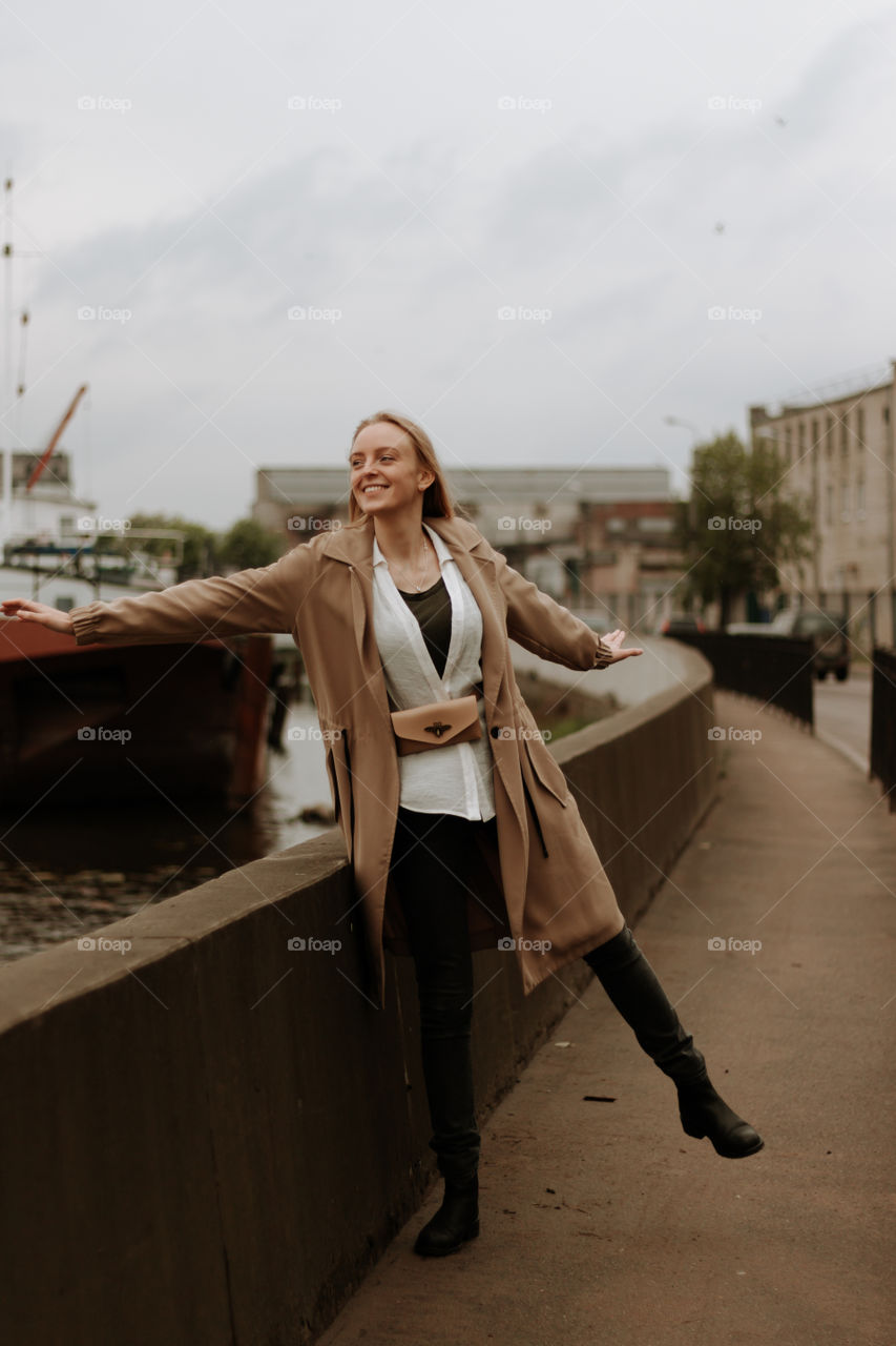 Blonde girl in a camel coat is having fun on a road to the city port. Street style outfit
