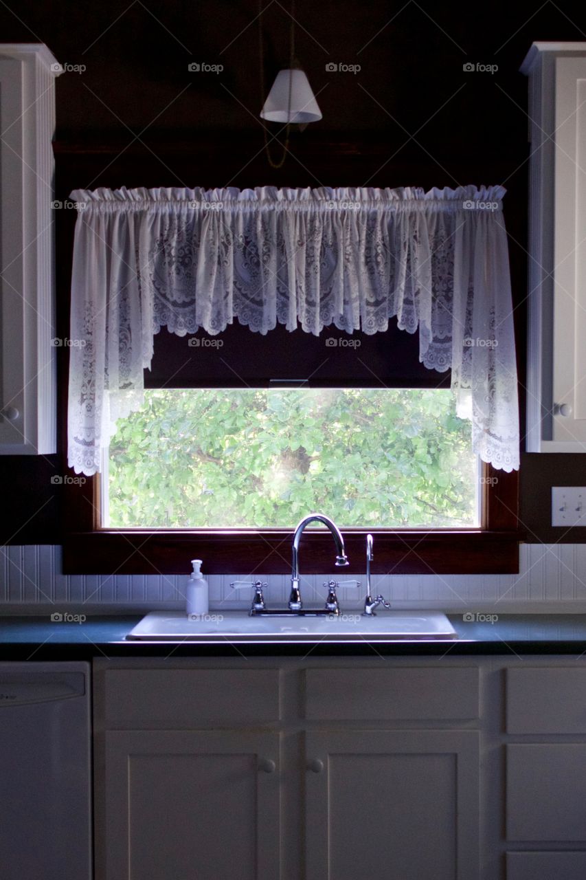 A white kitchen sink, vintage-style fixtures, open window, view of a leafy tree, soft sunlight through a white lace valance