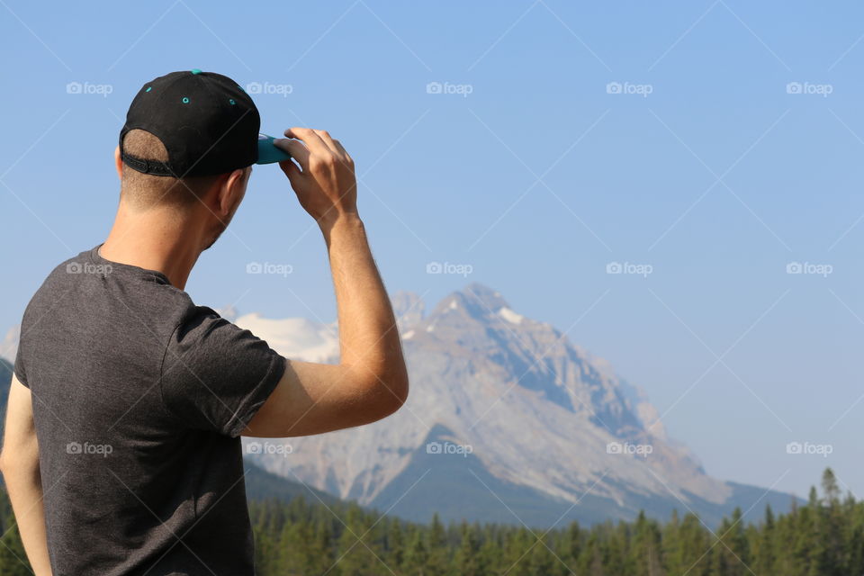 Young man wearing baseball cap, viewing and tipping hat brim to Canada's Rocky Mountains, 