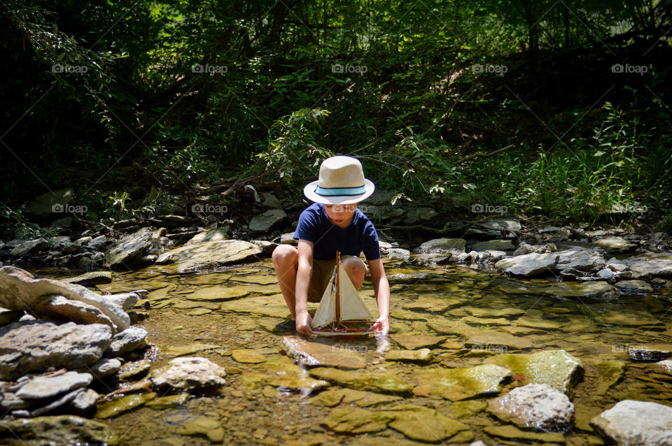 Boy playing with a toy sailboat in a creek during the summer