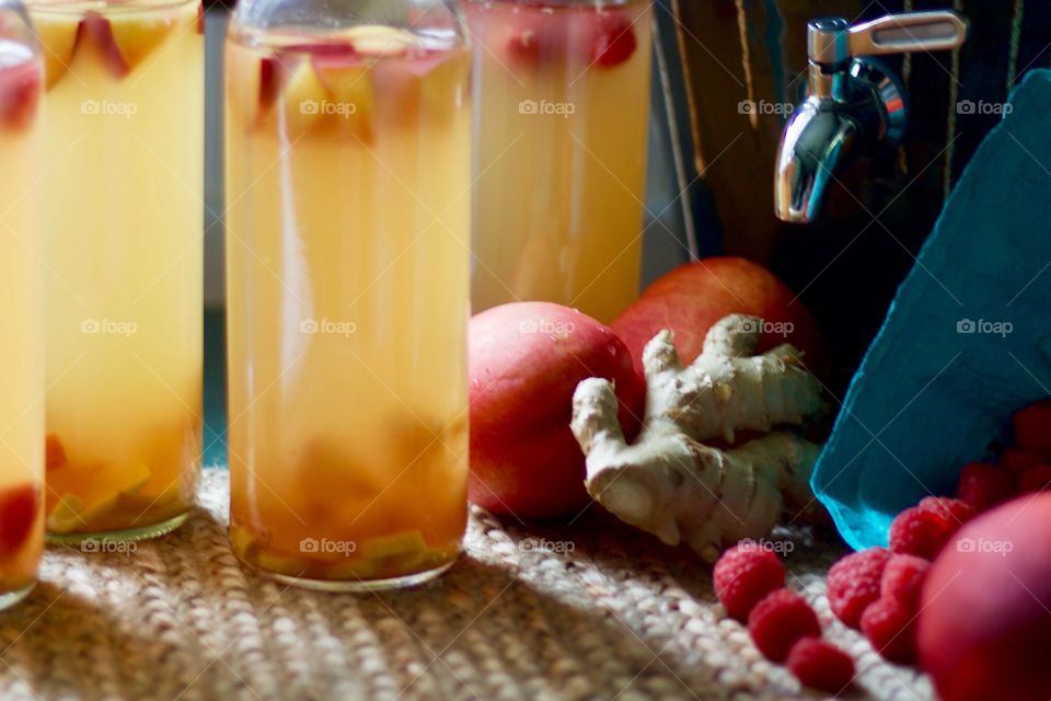 Kombucha, bottled for a second-ferment, flavored with nectarines, raspberries and ginger root slices on a natural fiber mat, whole nectarines and raspberries in a paper carton, stoneware kombucha crock with a stainless steel spigot in the background