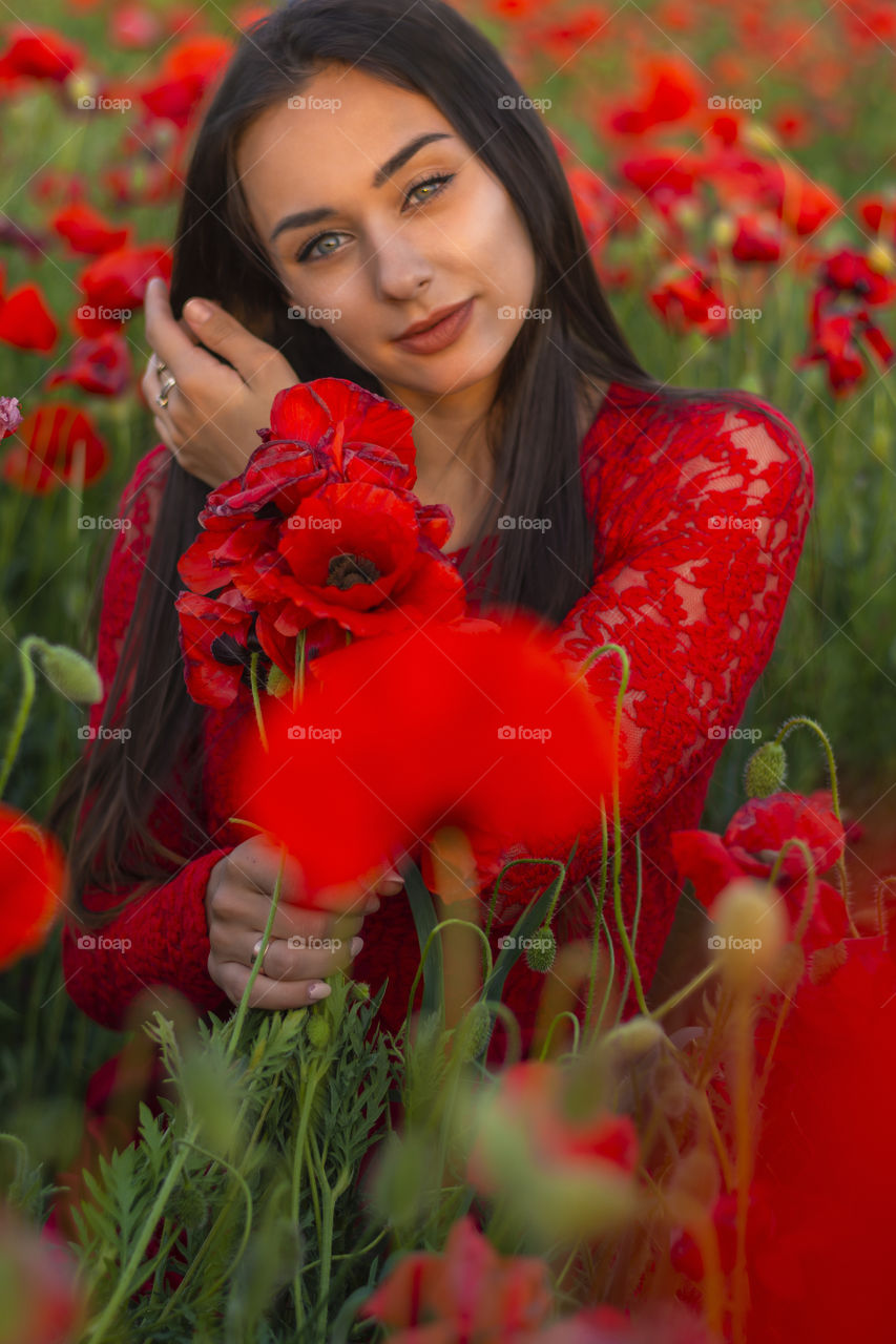 Close-up front view of a beautiful woman with a bouquet of poppies sitting in a poppy field and looking directly at the camera in nature