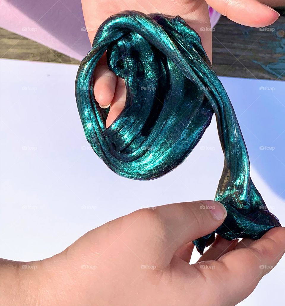 Turquoise Teal Metallic Color Slime Stretched And Held By A Child’s Two Hands With A White Background.