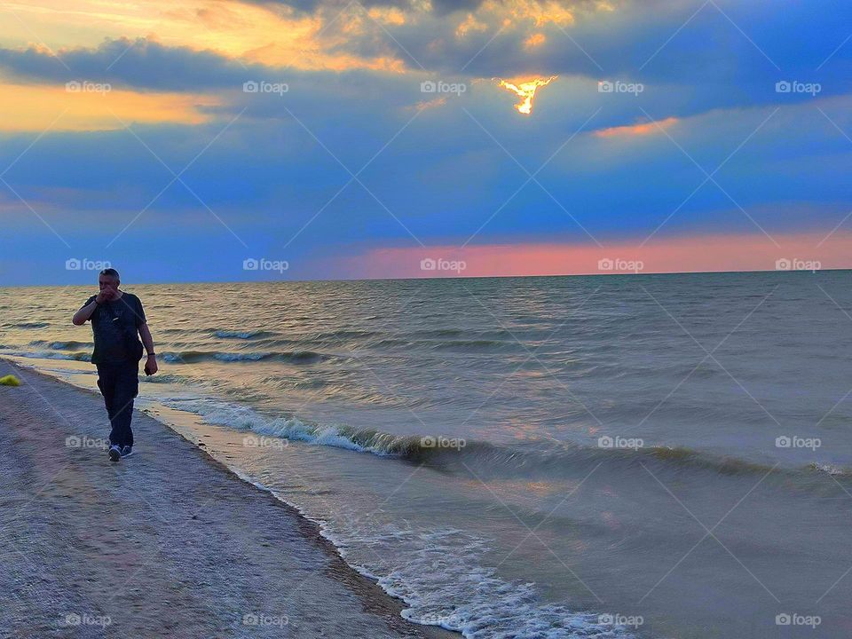 Beach coast. Summer evening. The shore of the azure sea with white waves against the backdrop of a summer sunset. The sky is painted yellow, blue and pink. A man walks along the beach
