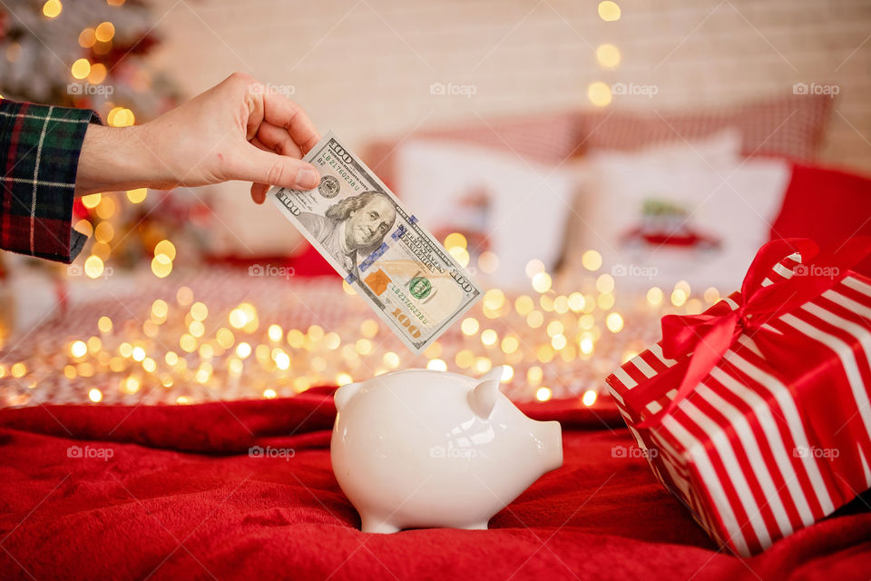 piggy bank with 100 dollars banknote in festive New Year atmosphere of scenery.  Magical bokeh with Christmas tree and bright lights.