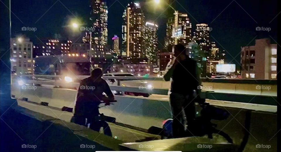 Two commuters appear only as faint dark silhouettes standing in the pedestrian lane of the “Pulaski Bridge” at night. They are currently waiting for the bridge to reopen for traffic. 2022. Hypnotic Productions