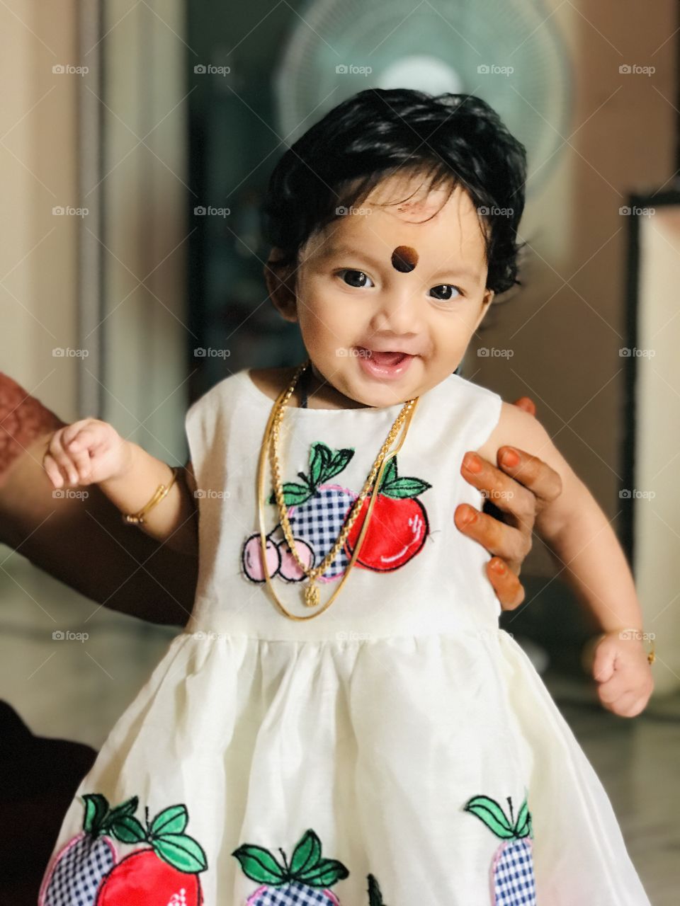 Indian little baby girl cute smile with cream colour dress and wearing neck chains and big Bindi on her forehead 