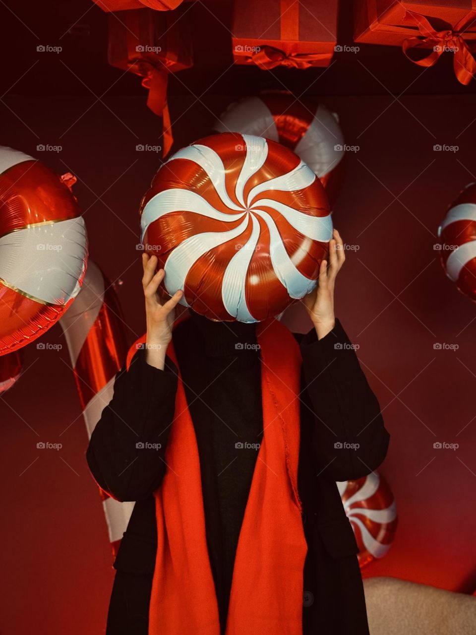 The image shows a person holding a large, red-and-white swirled candy-shaped balloon in front of their face. The scene is decorated with similar candy-themed props and red gift boxes.