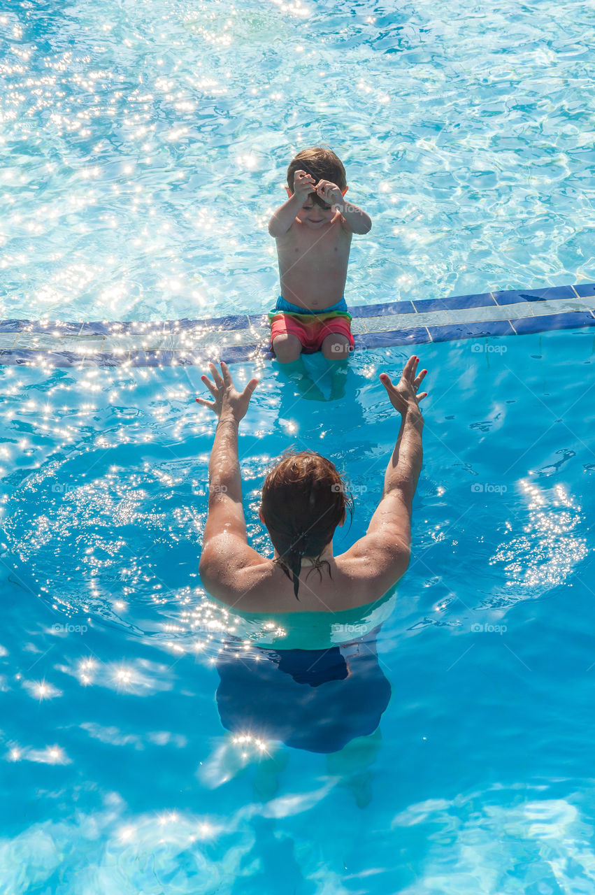 Swimming lesson. Mum encouraging her little son to get into water in swimming pool.